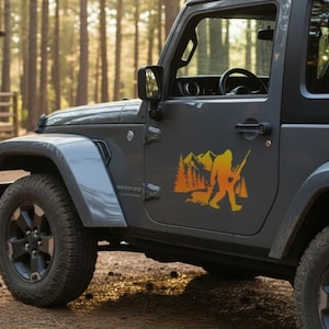 May include: A gray off-road vehicle featuring a yellow decal on the driver-side door. The decal depicts a silhouette of a large creature carrying a rifle, walking past a forest of pine trees and mountains, with a small animal near its feet.