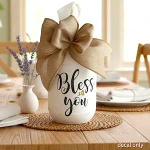 May include: A white cylindrical tissue holder featuring the black text "Bless you" on the front. The top is adorned with a large, tan burlap bow, and a white tissue is visible emerging from the center. The text "decal only" appears in the corner.