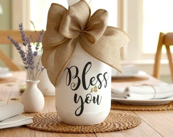May include: A white cylindrical tissue holder featuring the black text "Bless you" on the front. The top is adorned with a large, tan burlap bow, and a white tissue is visible emerging from the center. The text "decal only" appears in the corner.