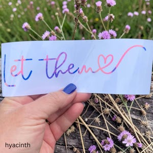 May include: A white rectangular decal featuring the text "let them" in a cursive font with a gradient of blue, purple, and pink colors. The letter "m" extends into a heart shape at the end of the phrase. The decal is held against a background.