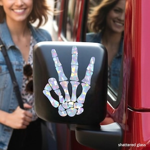 May include: Black side mirror with a holographic skeleton hand in a peace sign. The words "shattered glass" are in the bottom right corner. The mirror is attached to a red vehicle.