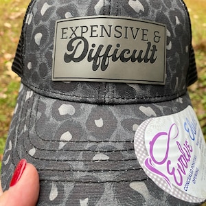 May include: A black and gray leopard print baseball cap featuring a rectangular patch with the text "EXPENSIVE & Difficult" in black lettering. The cap includes a white sticker on the brim that reads "Evolve" and mentions a concealed ponytail opening.
