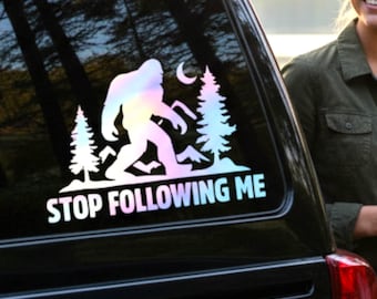 May include: An opal holographic vinyl decal applied to a vehicle window. The design features a silhouette of Bigfoot walking between two pine trees with a crescent moon and mountains in the background. The text STOP FOLLOWING ME is below.