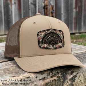 May include: A khaki baseball cap with a brown mesh back and a structured front panel. A rectangular patch on the front features a camouflage background with a black graphic illustration of a fanned-out turkey tail feather display.