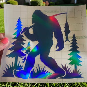 May include: A holographic vinyl decal featuring a silhouette of Bigfoot walking through a forest. The creature holds a fishing rod with a fish hanging from the line. The design includes two pine trees and grass, all rendered in a reflective, color-shifting material.