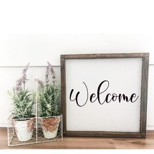 May include: A wooden framed sign with the word "Welcome" written in black cursive script. The sign is sitting on a wooden surface with two potted plants in a white wire basket.