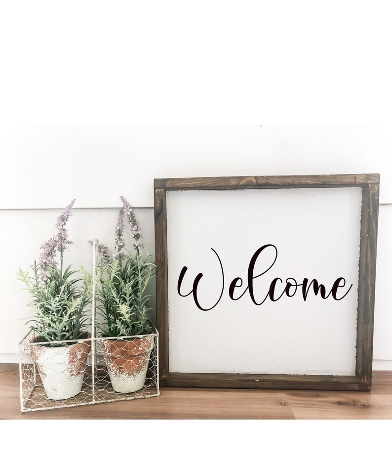 May include: A wooden framed sign with the word "Welcome" written in black cursive script. The sign is sitting on a wooden surface with two potted plants in a white wire basket.