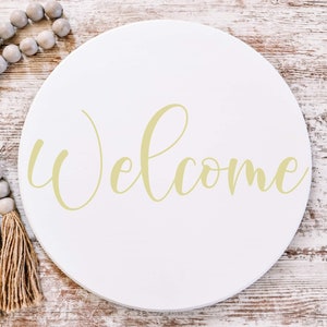 May include: A white round wooden sign with the word "Welcome" written in a cursive font in a light yellow color.