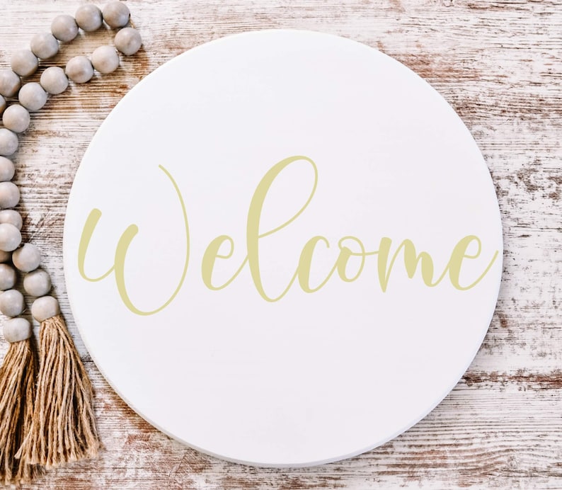 May include: A white round wooden sign with the word "Welcome" written in a cursive font in a light yellow color.