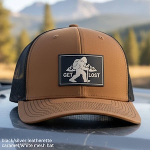 May include: A caramel-colored baseball cap with a black mesh back and a black rectangular patch on the front. The patch features a white graphic of a figure walking with a hiking stick in front of mountains and the text GET LOST in white letters.