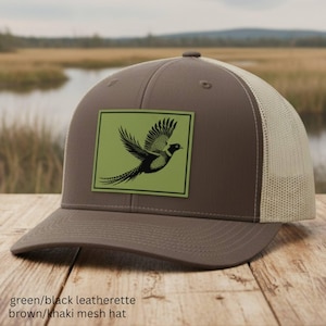 May include: A brown and khaki mesh hat featuring a rectangular green patch on the front. The patch displays a black graphic illustration of a pheasant in flight. The hat has a structured crown, a curved brim, and breathable mesh back panels.