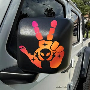 May include: Black side mirror cover with a colorful peace sign design. The design features a hand making the peace sign, an alien, and stars in shades of pink, orange, and yellow. The mirror is attached to a white vehicle.