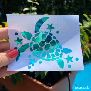 May include: A teal holographic vinyl decal featuring a stylized sea turtle surrounded by small stars and circular dots. The turtle shell is composed of multiple geometric shapes, and the entire design is set on a white rectangular backing paper.