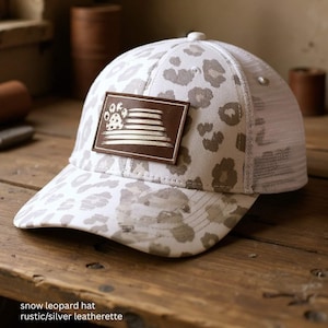 May include: A white and gray snow leopard print baseball cap with a mesh back. The cap features a brown leatherette patch with a paw print and striped design. The hat is on a wooden surface.