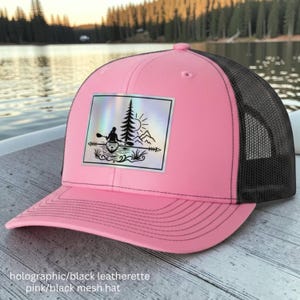 May include: A pink baseball cap with a black mesh back and a rectangular holographic patch on the front. The patch features a black graphic illustration of a person kayaking on water with a pine tree, mountains, and a sun in the background.