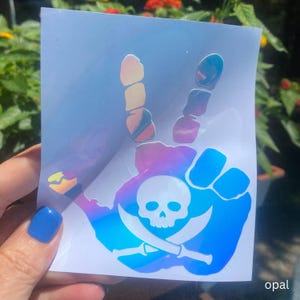 May include: An iridescent opal vinyl decal featuring a hand making a peace sign gesture. The design includes a white skull and crossbones graphic centered on the palm area. The sticker has a reflective, color-shifting surface that displays various hues.