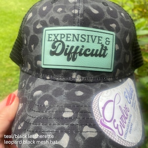 May include: A black and gray leopard print baseball cap with a black mesh back. A rectangular light green patch on the front features the black text "EXPENSIVE & Difficult". A white product label is attached to the lower right side.