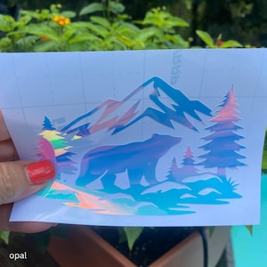 May include: An opal-colored vinyl decal featuring a silhouette of a bear standing in front of a mountain range and pine trees. The design has a reflective, iridescent finish that shifts colors, presented on a white grid-lined backing paper.