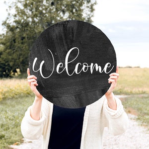 May include: A person holding a black round sign with the word "Welcome" written in white cursive lettering.