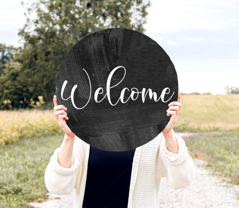 May include: A person holding a black round sign with the word "Welcome" written in white cursive lettering.