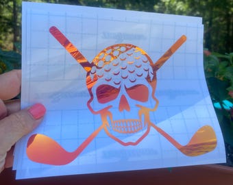 May include: A holographic sticker featuring a skull with a golf ball for a head and crossed golf clubs. The design is in shades of orange and red, with a reflective, iridescent finish. The text "orchid holo" is at the bottom.