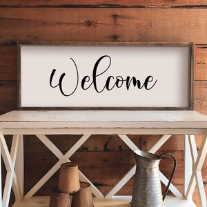 May include: A white wooden sign with a black cursive welcome message. The sign is hanging on a rustic wooden wall above a white wooden table with a metal pitcher and three brown ceramic pots.