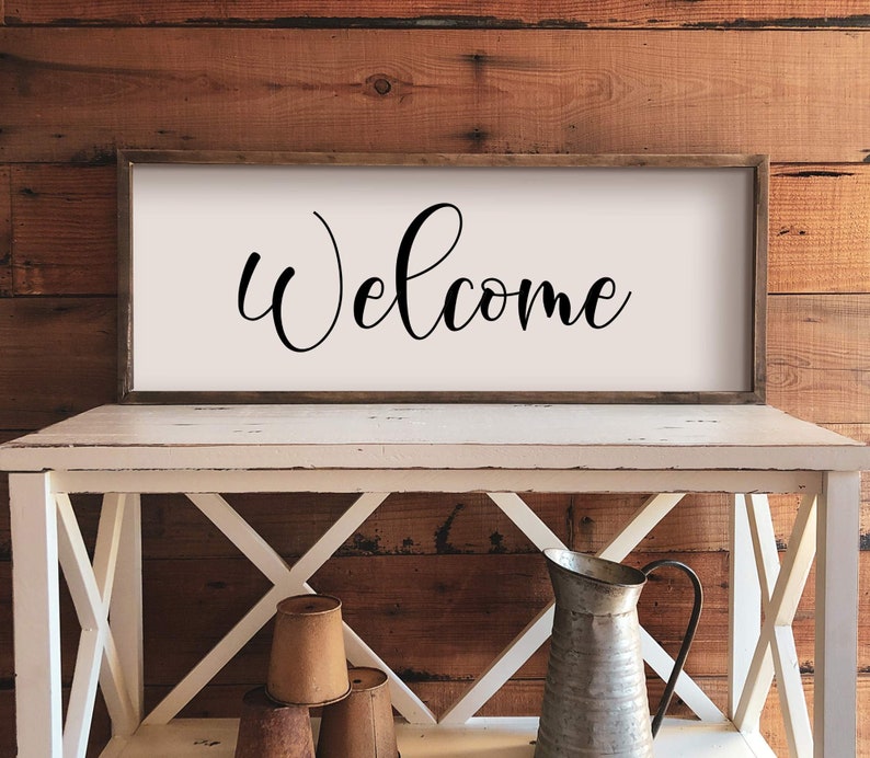 May include: A white wooden sign with a black cursive welcome message. The sign is hanging on a rustic wooden wall above a white wooden table with a metal pitcher and three brown ceramic pots.
