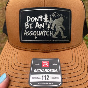 May include: A brown trucker hat with a black mesh back and a rectangular patch. The patch features the text "DON'T BE AN ASSQUATCH" with a Bigfoot silhouette and mountain graphic. A Richardson 112 Trucker tag is attached.