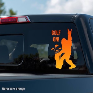 May include: A fluorescent orange vinyl decal applied to a vehicle rear window. The design features a silhouette of a creature resembling Bigfoot carrying a golf bag with clubs, with one hand raised in a rock-and-roll gesture and the text GOLF ON!