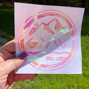 May include: A pink iridescent sticker with a mountain range and the word "G.A.V." inside a circle. The sticker has a holographic finish.