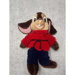 May include: A plush rabbit toy with brown fur, large ears, and a red shirt. The rabbit has a tan face with large eyes and a smiling expression. It is wearing blue pants and brown shoes.