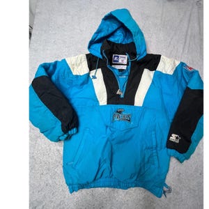 Puede incluir: Chaqueta anorak de los Carolina Panthers en turquesa, negro y blanco. La chaqueta tiene capucha, media cremallera y el logotipo de los Panthers en la parte delantera. El logotipo de Starter es visible en la manga. Ropa deportiva vintage.