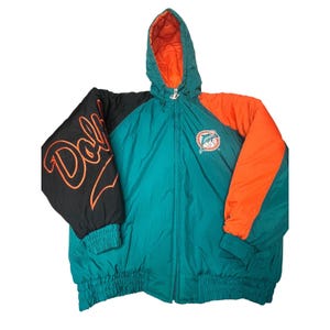 Könnte beinhalten: Eine türkisfarbene, schwarze und orangefarbene Kapuzenjacke mit dem Miami Dolphins Logo auf der Brust. Der rechte Ärmel zeigt das Wort Dolphins in orangefarbener Schreibschrift. Die Jacke hat einen durchgehenden Reißverschluss, gerippte Bündchen und ein gestepptes orangefarbenes Innenfutter.