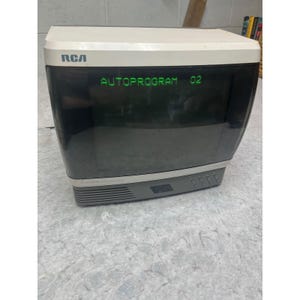May include: A vintage RCA television set with a beige top casing and a dark gray base. The screen displays the green text AUTOPROGRAM 02. The front panel features a series of small control buttons and a speaker grille located below the screen.