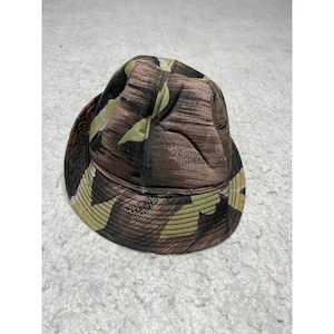 May include: A camouflage bucket hat in shades of brown, green, and black. The hat features a rounded crown and a downward-sloping brim. The North Face logo is visible on the side of the hat.