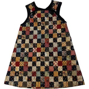 Könnte beinhalten: Ein ärmelloses Trägerkleid mit einem Patchwork-Design aus kleinen Quadraten in verschiedenen Mustern und Farben, darunter Rot, Marineblau, Hellbraun und Creme. Das Kleidungsstück hat einen schwarzen Saum am Ausschnitt und an den Ärmelausschnitten mit zwei schwarzen Knöpfen.