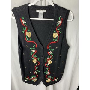 May include: Black button-up sweater vest with a V-neck. The vest is decorated with embroidered gold bells, red ribbons, green leaves, and white flowers. The vest is hanging on a white hanger.