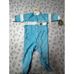 May include: A light blue and white long-sleeved footed sleeper made of a textured, plush fabric. The garment features a horizontal white stripe across the chest, a front button closure, and a small embroidered graphic with text on the upper left side.