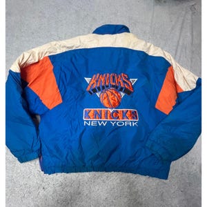 May include: Blue, orange, and cream-colored vintage Knicks jacket. The jacket features the Knicks logo with a basketball in the center. The words "Knicks" and "New York" are embroidered on the jacket.