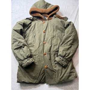 Puede incluir: Un abrigo con capucha verde oliva con una capucha forrada de pelo marrón. El abrigo tiene una botonadura frontal, dos bolsillos delanteros y puños ajustables. El abrigo está hecho de un material duradero, perfecto para el frío.