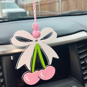 Puede incluir: Un amuleto de coche rosa y blanco con un lazo y dos cerezas. El amuleto está hecho de acrílico y tiene un tallo verde.
