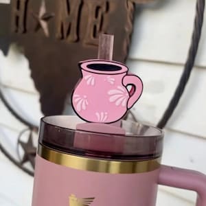 以下が含まれることがあります： ピンク色のタンブラー。ピンク色の蓋とハンドルが付いています。白い花の模様が入ったピンク色のマグカップ型のストローのトッパーが蓋に付いています。タンブラーの上部には金のバンドと金のロゴが付いています。