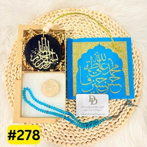 May include: A gift set comprising a black circular ornament with gold calligraphy, a blue book with gold accents and calligraphy, light blue prayer beads, and a small round stone. Presented in a box on a woven mat.