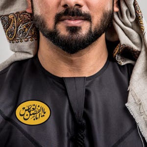 Puede incluir: Un thobe negro con un emblema amarillo y negro con escritura árabe. El thobe tiene un cuello mandarín y una borla decorativa. Una bufanda estampada en beige y marrón está drapeada sobre la cabeza.