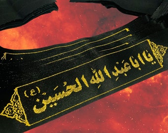 May include: Black fabric with gold Arabic script. The text reads "Ya Aba Abdillah Al-Hussein". The fabric features a decorative border and is set against a red and orange background, reminiscent of a nebula.
