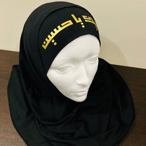 May include: A black hijab with a gold Arabic inscription on the forehead band.