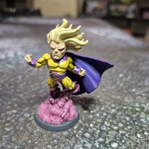 May include: A miniature figurine of a superhero in a dynamic pose. The figure is predominantly yellow with purple accents, a purple cape, and flowing blonde hair. It stands on a pink cloud base, mounted on a gray circular platform.