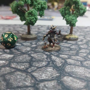 Op de afbeelding: Een groene 20-zijdige dobbelsteen met gouden nummers, een miniatuurfiguur van een katachtig wezen met een hoed en een zwaard, en twee kleine groene bomen op een grijs stenen oppervlak.