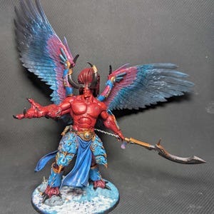 Peut inclure: Figurine miniature détaillée d'une créature démoniaque à la peau rouge, avec de grandes ailes bleues et violettes et des cornes. La figurine tient une lame incurvée et se tient sur une base enneigée. La figurine est peinte avec des détails complexes.