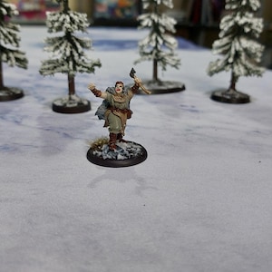 May include: A miniature figure of a person wearing a brown tunic and holding an axe. The figure is standing on a round base with a snowy landscape background.